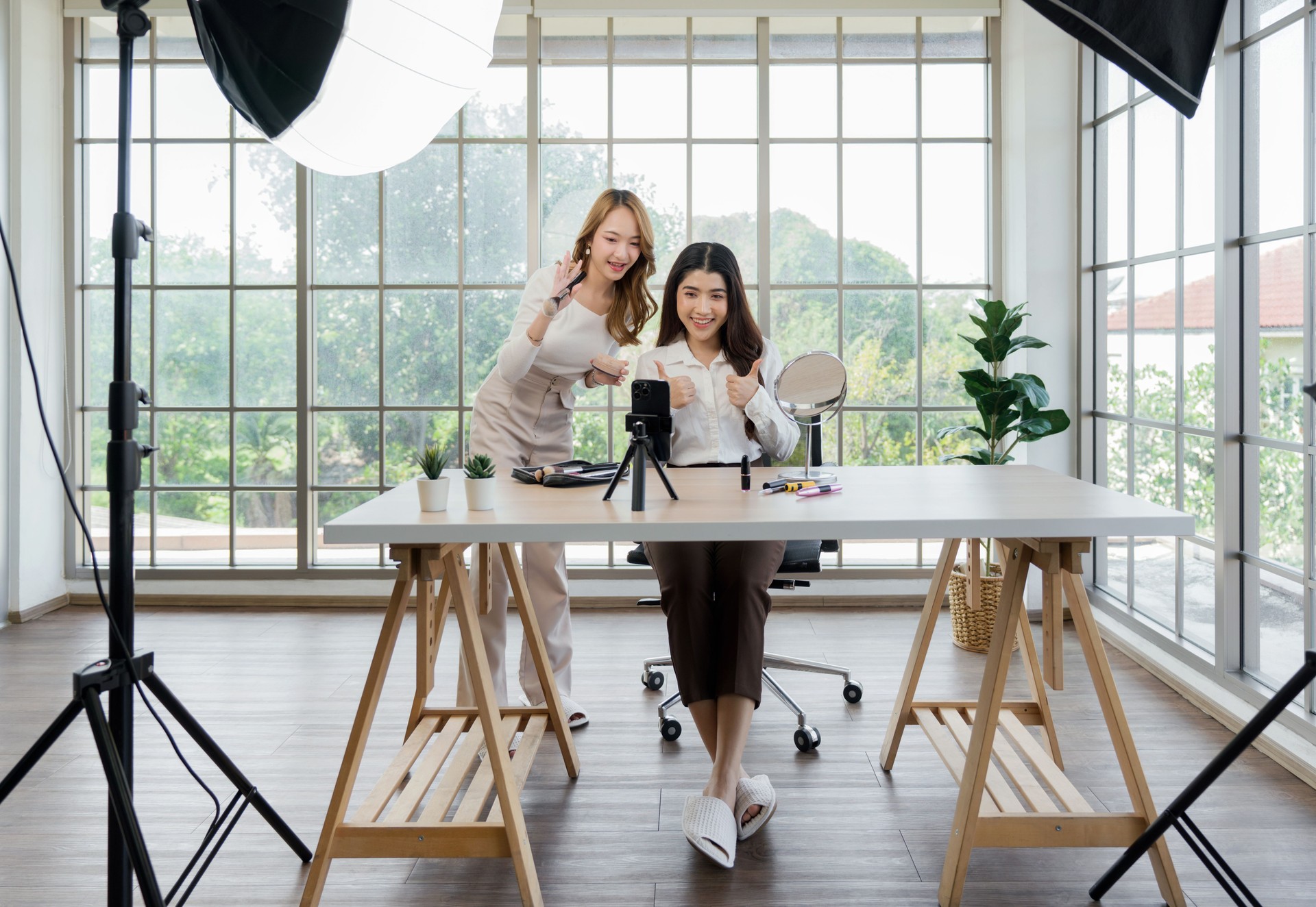 Two asian women creating content for social media. They are using a studio setup that include a smartphone mounted on a tripod, lighting equipment and various makeup products arrayed on the table.